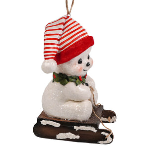 A glittery white snowman ornament with a red striped hat and holly collar on a brown sleigh by bethany lowe design