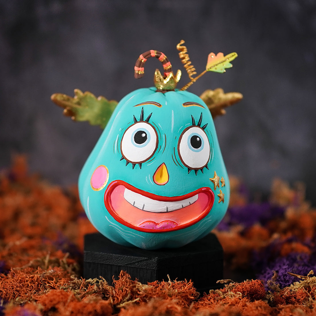 A turquoise pixie pumpkin figurine with oversized cartoon eyes, a wide smile, and golden leafy antennae