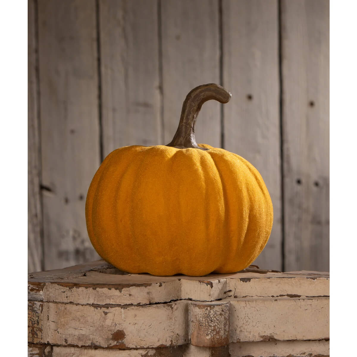 A bethany lowe flocked mustard jol plush orange pumpkin with textured surface and dark brown stem on white bricks