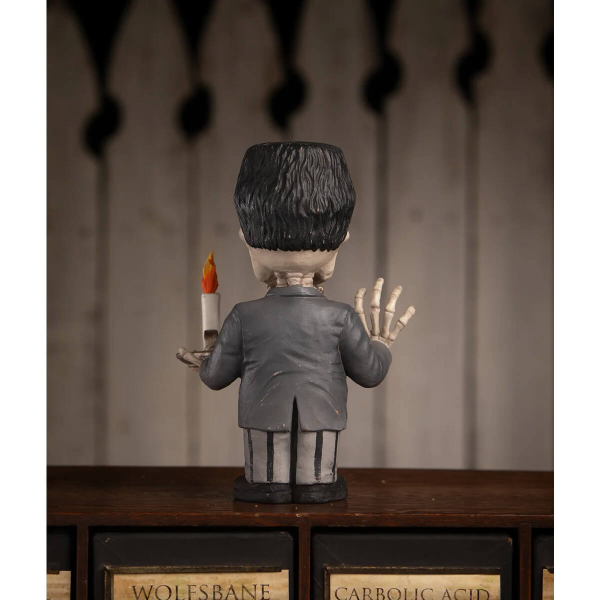 A gray franken skelly figurine with black hair in a formal suit holding a lit candle with skeletal hands