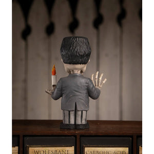 A gray franken skelly figurine with black hair in a formal suit holding a lit candle with skeletal hands