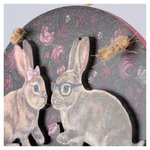 Circular wooden valentine’s day ornament with two illustrated rabbits on a dark rose patterned background