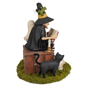 Black witch figurine with tall hat and cape reading a book on wooden trunk perfect for halloween witches decor and ghost stories