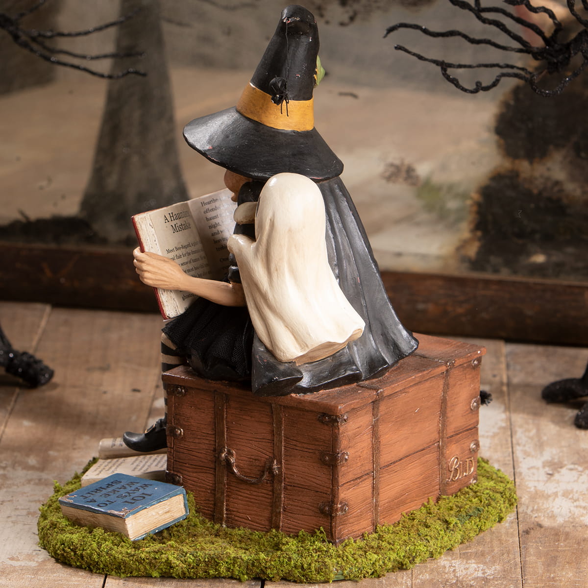 A halloween witches figurine with black hat and cape sitting on a wooden trunk reading a book for ghost stories decor