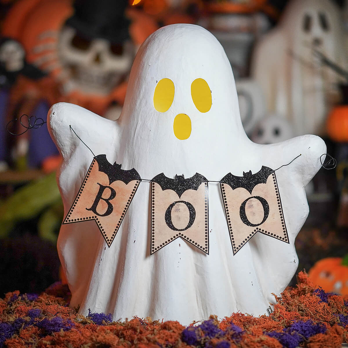 White ghost figurine with yellow eyes holding a boo garland of black glitter bats on beige pennants for halloween ghosts decor