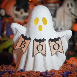 White ghost figurine with yellow eyes holding a boo garland of black glitter bats on beige pennants for halloween ghosts decor