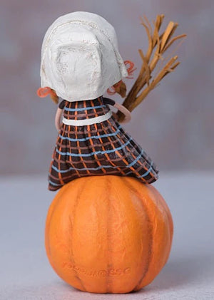 A lori mitchell grateful greta figurine in a white bonnet and plaid dress holding a broom on an orange pumpkin