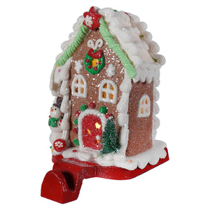 A glittery brown led lighted gingerbread candy house ornament with white icing, red door, snowman, and green wreath