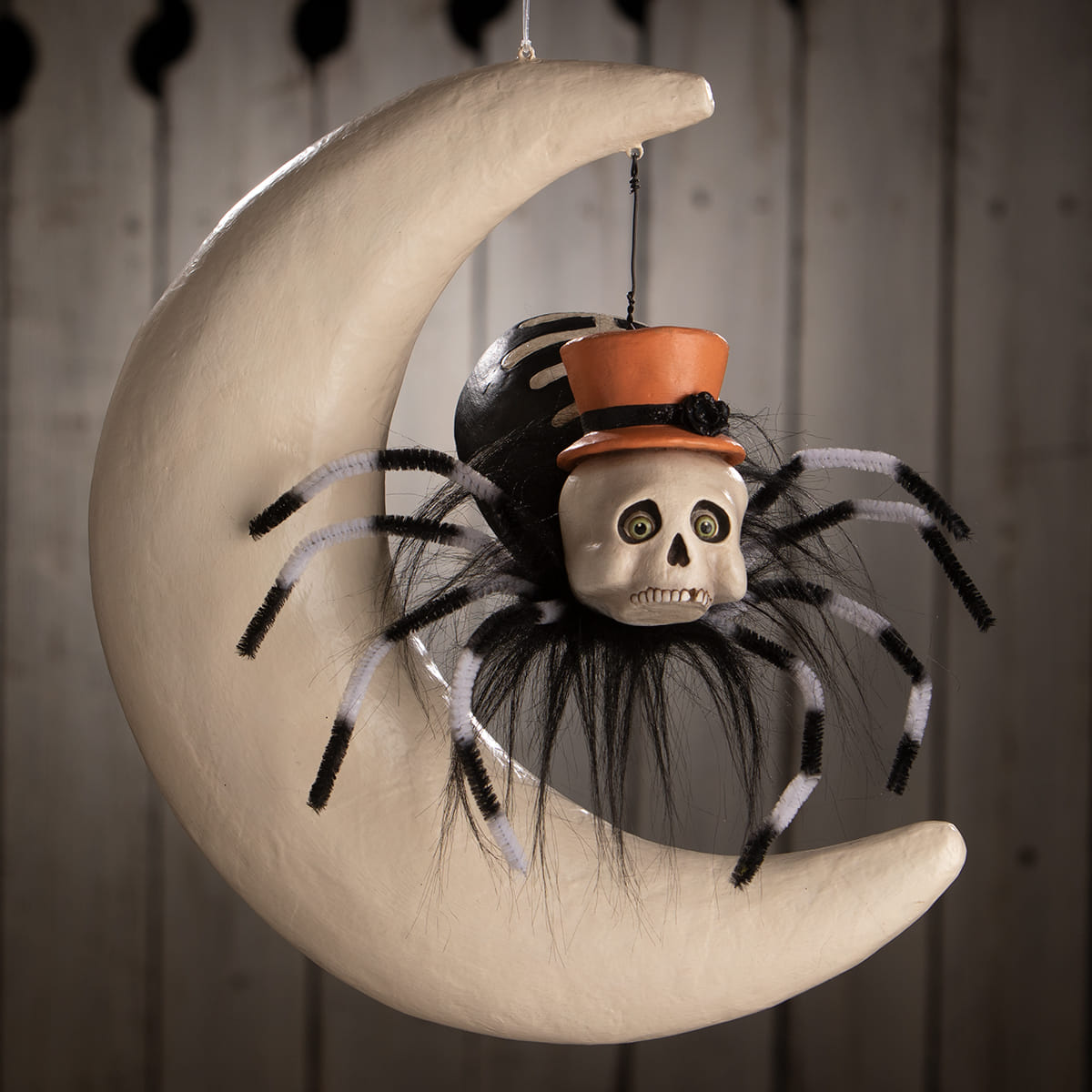 A whimsical hanging spider on moon decoration with black and white pipe cleaner legs and an orange top hat