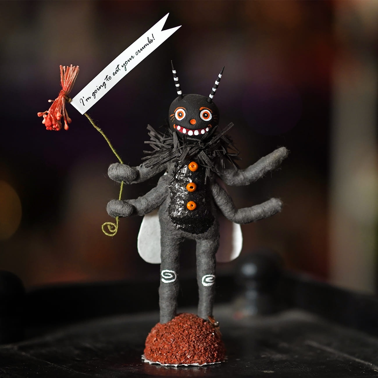 A whimsical black felt happy fly figure with orange button eyes holding a red flower and crumb-eating banner