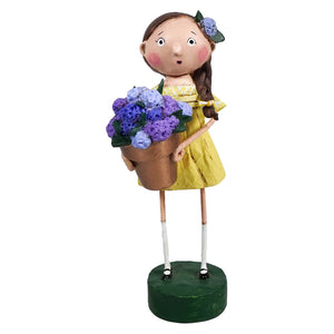 Whimsical decorative figurine of a girl in a yellow dress holding purple flowers, mounted on a green base.