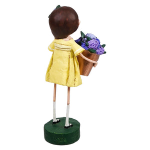 Decorative figurine wearing a yellow dress and holding a pot of purple flowers, mounted on a green base.