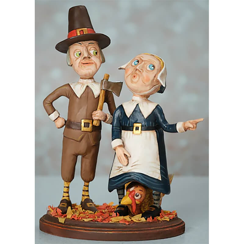 Clay or ceramic figurine of a Pilgrim couple with a turkey at their feet standing among autumn leaves.