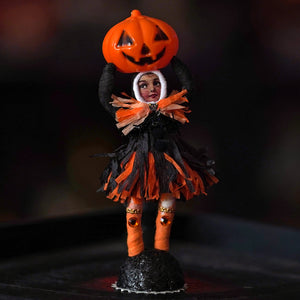 A miniature halloween heavy jack o lantern figure in black and orange fringed outfit holding an orange jack o lantern on its head