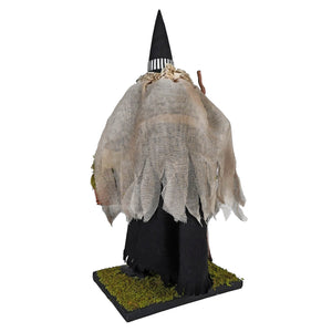 A spooky hildegard witch figurine with a black striped hat and tattered beige fabric holding a potion basket on mossy base