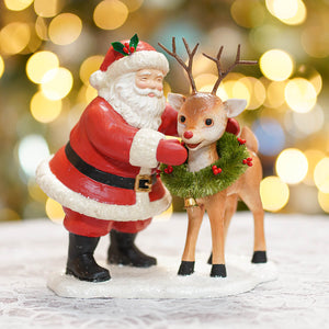 Red and white santa figures gently hugging a tan reindeer with wreath in folk art santas style santa with rudolph