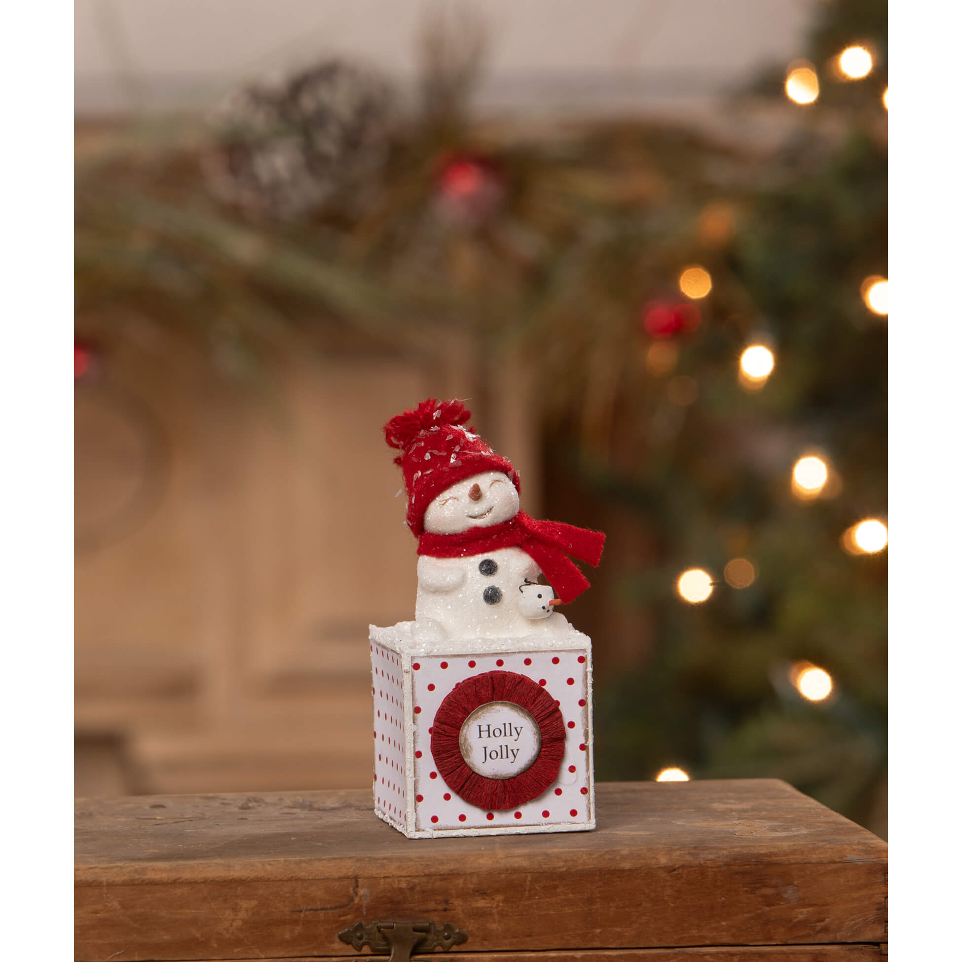A white ceramic holly jolly snowgirl snowman figure wearing red knit hat and scarf sits on festive polka dot box