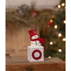 A white ceramic holly jolly snowgirl snowman figure wearing red knit hat and scarf sits on festive polka dot box