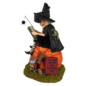 A whimsical bethany lowe halloween witch figurine with black pointed hat reading a book on an orange pumpkin base with black cat theme