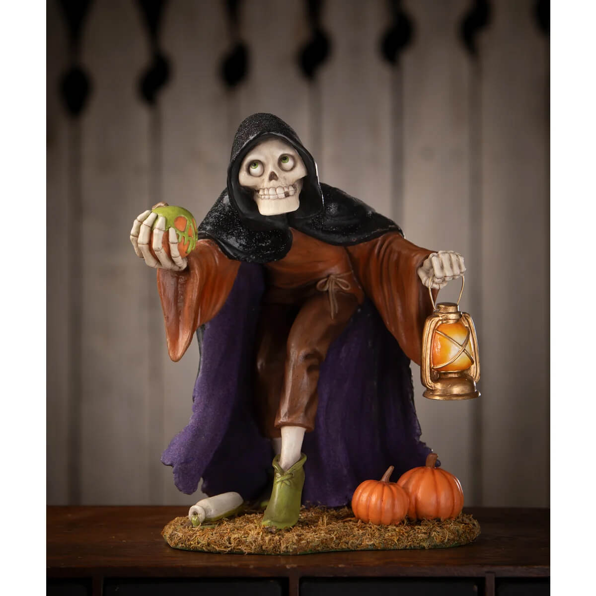 A halloween-themed igor skeleton figurine in black hooded cloak holding an apple and lantern with pumpkins and green boots