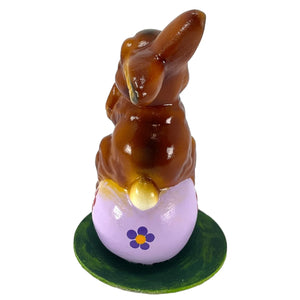 A glossy brown chocolate schaller easter bunny paper mache perched on a lavender egg with purple flowers on green base