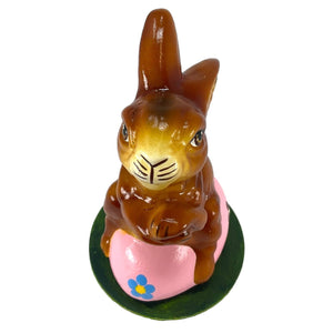 A glossy brown ino schaller easter rabbit hand-painted paper mache sitting on a pink egg with a blue flower on green base
