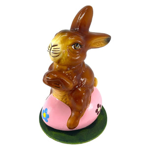 A glossy brown ino schaller easter paper mache bunny sits on a pink egg with blue and purple flowers