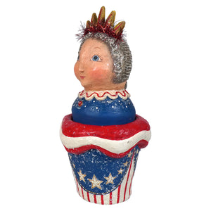A whimsical ceramic johanna parker lady liberty container shaped like a red white and blue cupcake with a crowned girl inside