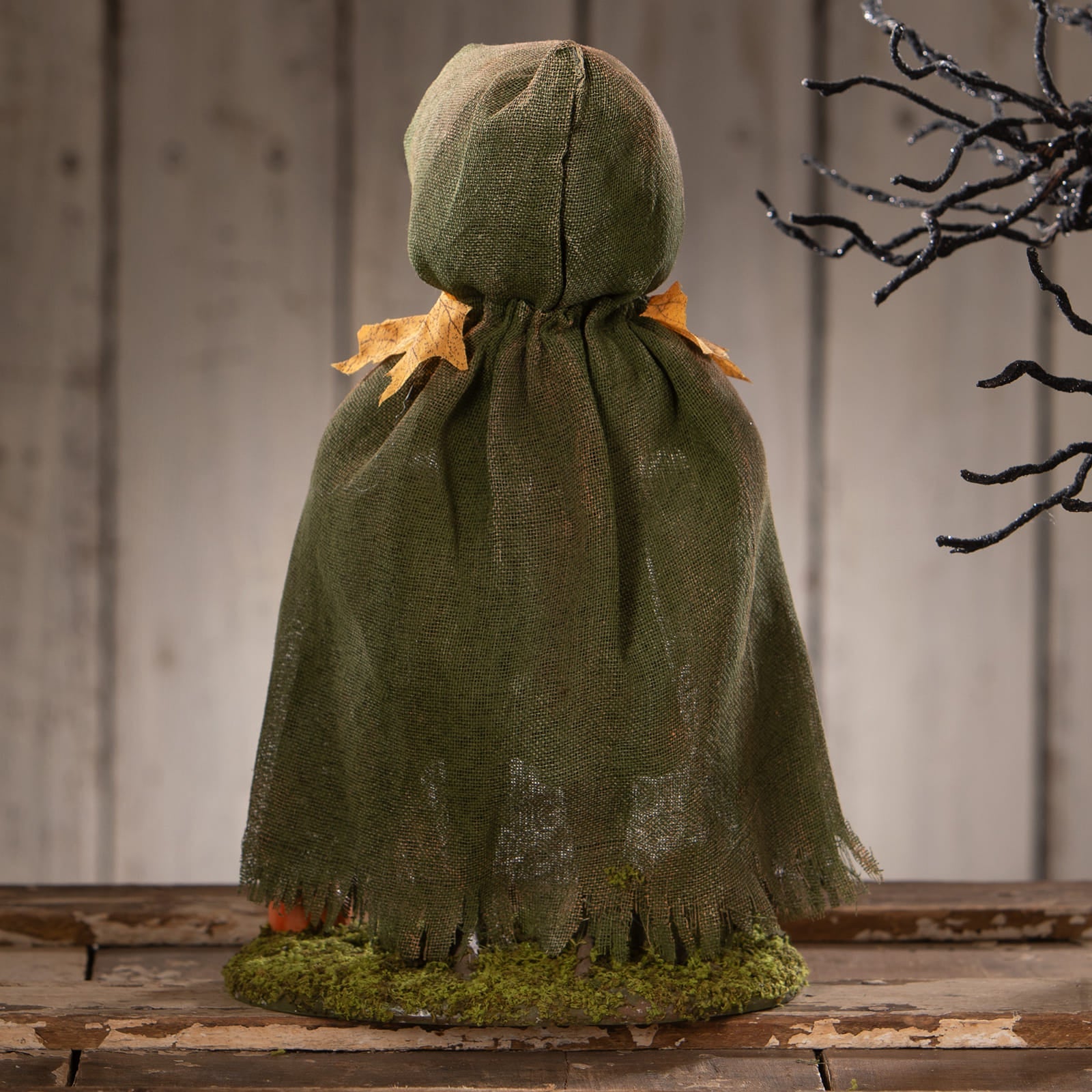 A rustic olive green burlap hooded cloak with frayed edges and dried orange leaves on a mossy base for halloween figural decor pumpkin