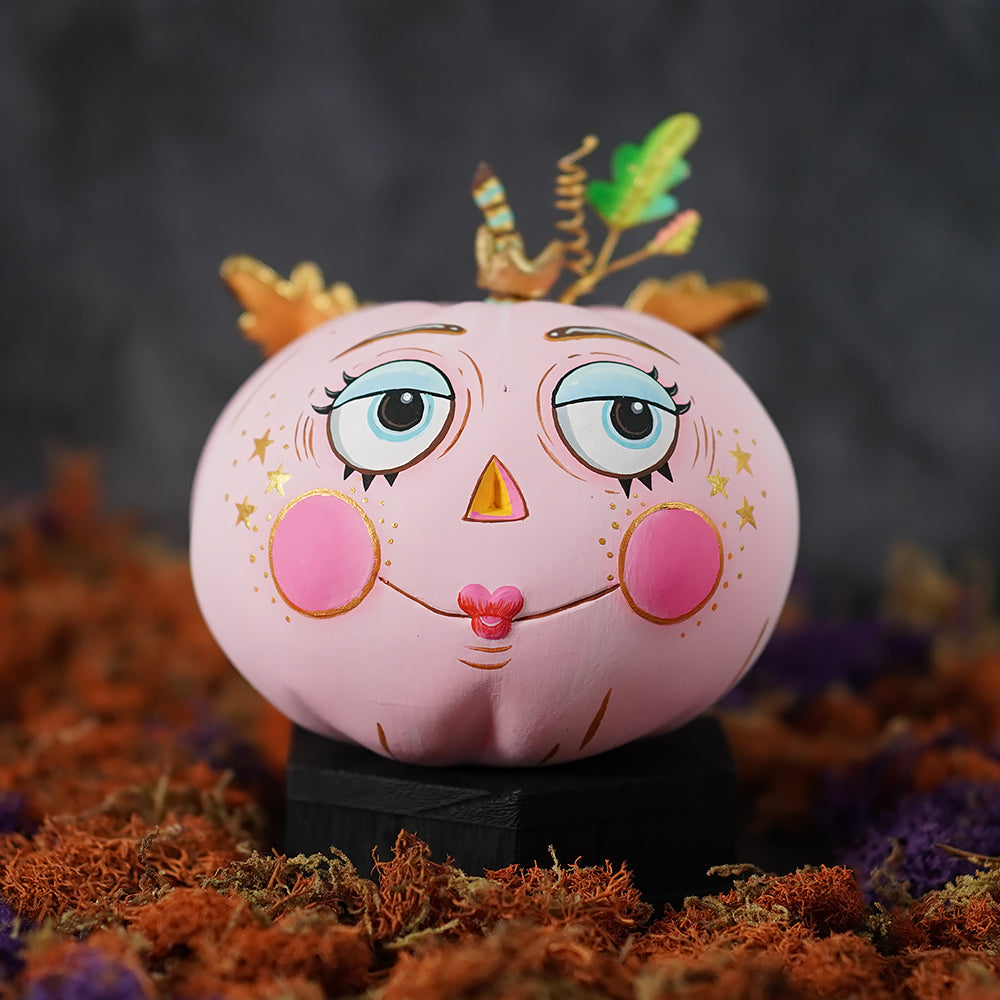 A pink pixie pumpkin with expressive blue eyes rosy cheeks gold stars and a whimsical antler crown for halloween pumpkin display