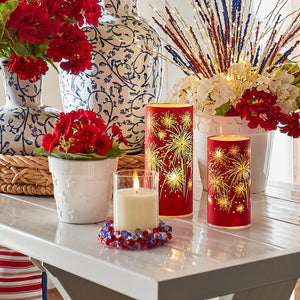 Red decorative candle holders with gold firework designs alongside white candles and red geranium flowers.