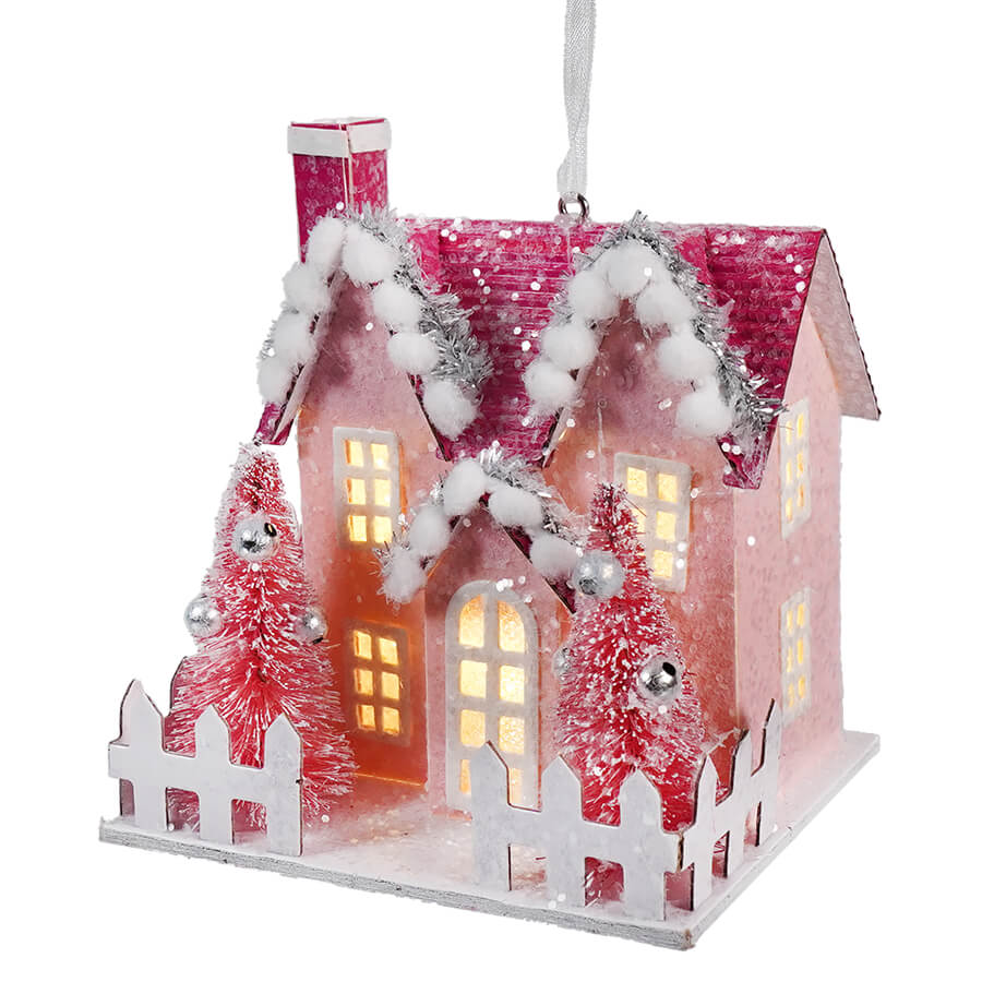 Illuminated pink Christmas house ornament with snowy roof and miniature trees.