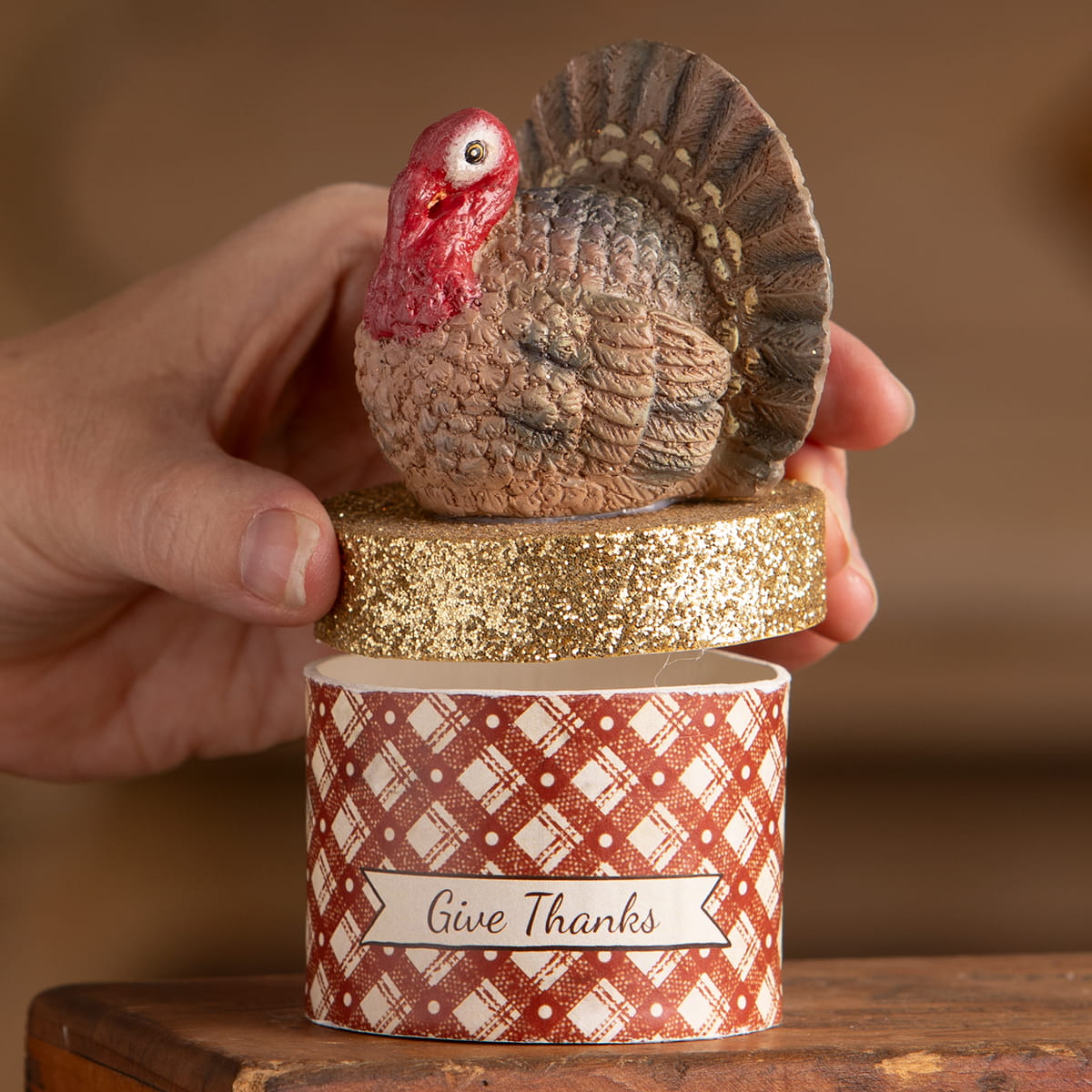 A brown textured turkey figurine with a red head sits on a glittery gold lid of a red plaid thanksgiving turkeys box