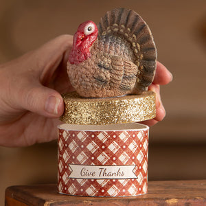 A brown textured turkey figurine with a red head sits on a glittery gold lid of a red plaid thanksgiving turkeys box