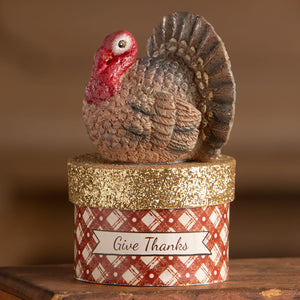 A ceramic little turkey on box with a red head and brown body sitting on a glittery gold plaid thanksgiving decor box