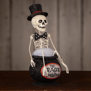 A white skeleton decor figure wearing a black glitter top hat and bow tie sitting in a magic cauldron ornament