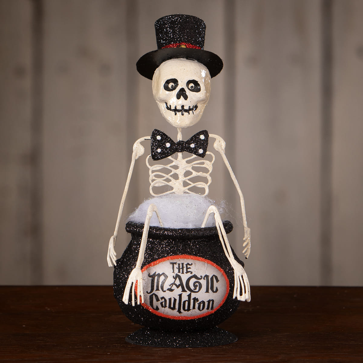 A white skeleton decor figure wearing a black glitter top hat and bow tie seated inside a black magic cauldron ornament