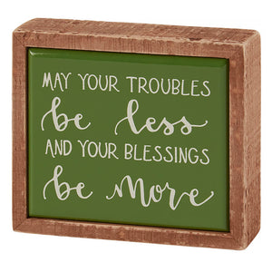 A wooden-framed green sign displaying an inspirational message about troubles and blessings.