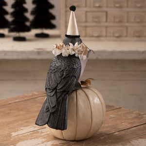 A black messenger crow figurine with detailed feathers wearing a white party hat and floral crown on an off-white pumpkin from bethany lowe halloween
