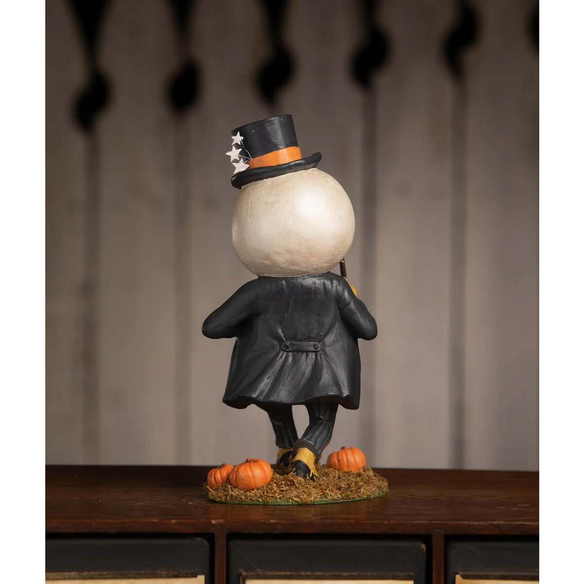 A whimsical harvey moon man pumpkin figure in a black starry top hat and glossy black coat standing on hay with pumpkins