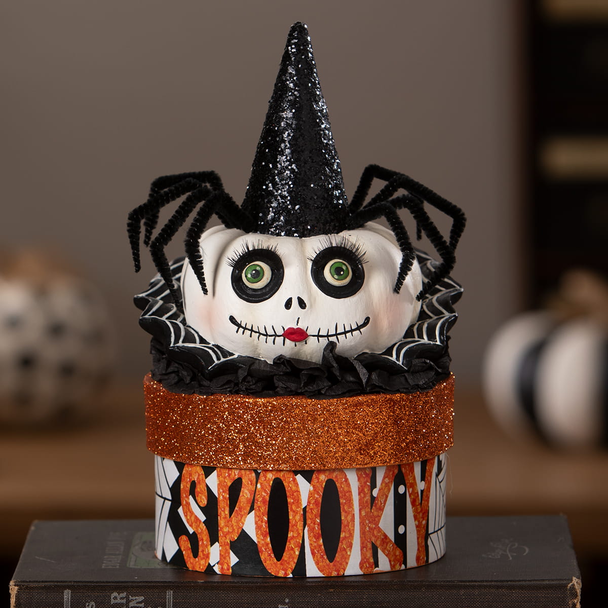 A white skull with green eyes and red mouth wearing a black glittery cone hat surrounded by black pipe cleaner spiders on mrs casperita box