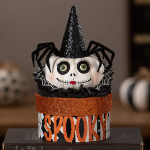 A white skull with green eyes and red mouth wearing a black glittery cone hat surrounded by black pipe cleaner spiders on mrs casperita box