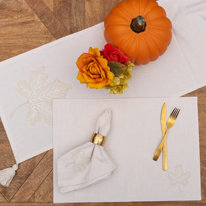 Place setting with a white placemat, gold utensils, and a napkin in a gold ring, accompanied by a pumpkin and flowers.