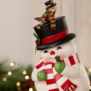 A white bethany lowe designs christmas snowman figure with black top hat, red ribbon, striped scarf, and green gloves holding a pipe