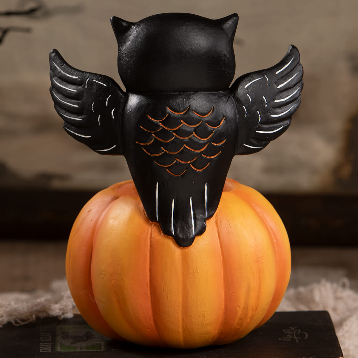 Black owl figurine with white-lined wings and orange-scaled back perched on an orange halloween pumpkin
