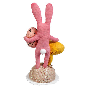 Pink bunny child handcrafted folk art figurine holding speckled egg on glittery base with yellow chick detail