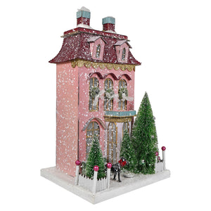 A festive pink chateau house ornament with glittery red roof gold trim and miniature snowy green trees for a village display