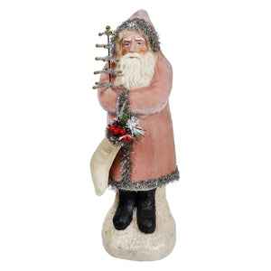 A vintage velvet belsnickle figurine in a pink coat with silver tinsel trim holding a tree on a snowy base with stocking