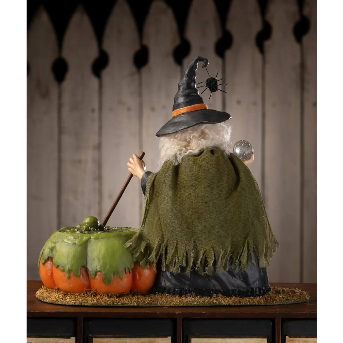 A poison brewing pruella witch figurine with a black hat, green fringed cloak, and pumpkin cauldron accessory
