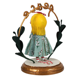 A whimsical praying for peace figurine of a girl with yellow hair in a blue floral dress under a golden black-leaf arch perfect as a child’s christmas figure